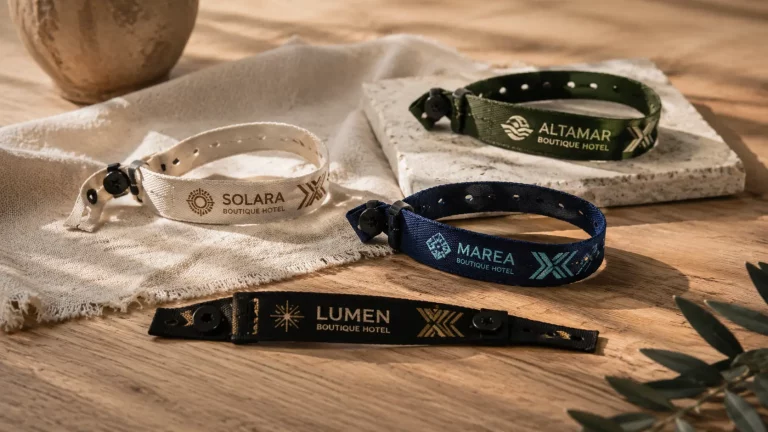 Cuatro pulseras de tela ajustables personalizadas con logotipos de hoteles boutique, en colores blanco, verde, azul y negro, dispuestas sobre una mesa de madera con un paño de tela y una base de piedra. La escena está iluminada con luz natural y presenta una decoración de estilo cálido y minimalista.